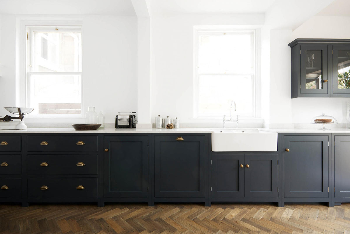 A bright and simple pantry-blue Shaker galley kitchen features a long sink and cooker run with a double Belfast sink, an antique silver Ionian tap, and glazed wall cupboards. Beautifully decorated with fundamental kitchen equipment such as a silver toaster, matching silver coffee, tea, and sugar containers, a glass cake stand and dome, a vintage scale, and reclaimed wooden parquet, it creates a stylish and effortless space.
