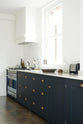An oblique view of a bright and simple pantry-blue Shaker galley kitchen features a long sink and cooker run with a silver gas cooker and a Shaker canopy. Beautifully decorated with fundamental kitchen equipment such as a silver toaster, a vintage scale, and reclaimed wooden parquet, it creates a stylish and effortless space.