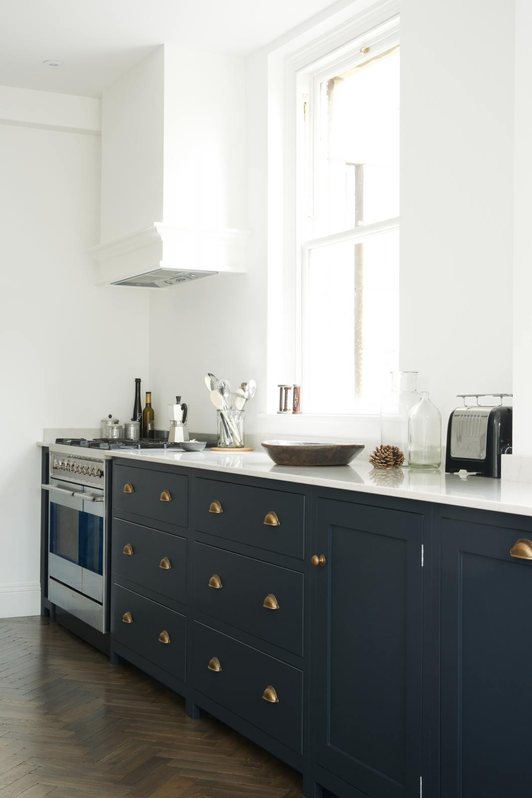 An oblique view of a bright and simple pantry-blue Shaker galley kitchen features a long sink and cooker run with a silver gas cooker and a Shaker canopy. Beautifully decorated with fundamental kitchen equipment such as a silver toaster, a vintage scale, and reclaimed wooden parquet, it creates a stylish and effortless space.