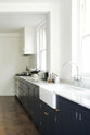 An oblique view of a bright and simple pantry-blue Shaker galley kitchen features a long sink and cooker run with a double Belfast sink, an antique silver Ionian tap, silver gas cooker, and a Shaker canopy. Beautifully decorated with fundamental kitchen equipment such as a silver toaster, matching silver coffee, tea, and sugar containers, a vintage scale, and reclaimed wooden parquet, it creates a stylish and effortless space.