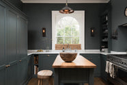 A charcoal-black, classic shaker kitchen features a U-shaped sink and cooker run with Carrara marble worktop, a chrome Ionian tap, open countertop cupboards, a prep table with a solid oak worktop and a bank of fitted pantry cabinets. Beautifully decorated with a large vintage ceramic bowl, steampunk wall lights, a copper pendant light, fresh pears, and the original Georgian wooden sliders, the family kitchen strikes a wonderful balance between modern style and timeless character.