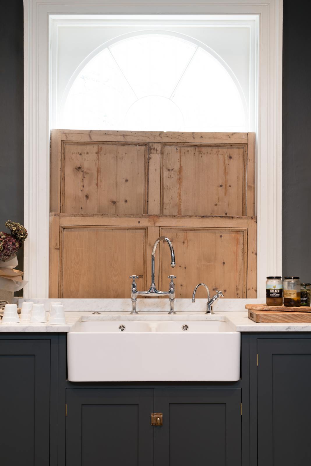 A charcoal-black, classic shaker kitchen features a U-shaped sink and cooker run with Carrara marble worktop, a double Belfast sink, and a chrome Ionian tap. Beautifully decorated with jars of different pickles, a vase of flowers, plastic cups, and the original Georgian wooden sliders, the family kitchen strikes a wonderful balance between modern style and timeless character.