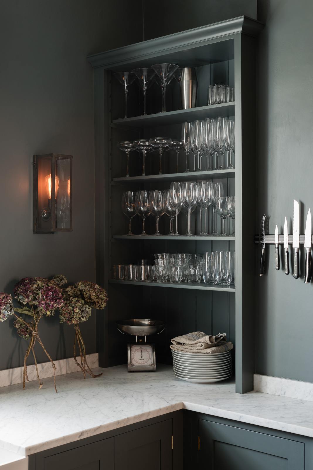 A quiet corner of a charcoal-black, classic shaker kitchen features a U-shaped sink and cooker run with Carrara marble worktop, a double Belfast sink, chrome Ionian tap and open countertop cupboards. Beautifully decorated with a magnetic kitchen knife holder, a steampunk wall light, a steel weight, branches of flowers, and the original Georgian wooden sliders, the family kitchen strikes a wonderful balance between modern style and timeless character.