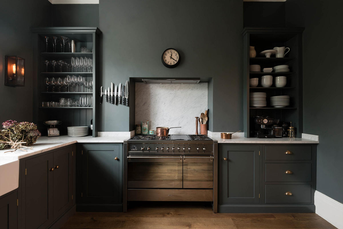 A charcoal-black, classic shaker kitchen features a U-shaped sink and cooker run with Carrara marble worktop and splashback, a double Belfast sink, and a silver gas stove. Beautifully decorated with a magnetic kitchen knife holder, a steampunk wall light, a steel weight, and branches of purple and green flowers, the family kitchen strikes a wonderful balance between modern style and timeless character.