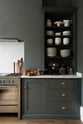 A quiet corner of a charcoal-black classic Shaker kitchen features a U-shaped sink and cooker run with a Carrara marble worktop, matching splashback, and open countertop cupboards. Beautifully decorated with copper pans, simple white ceramic plates and bowls, and a black espresso maker, the family kitchen strikes a wonderful balance between modern style and timeless character.