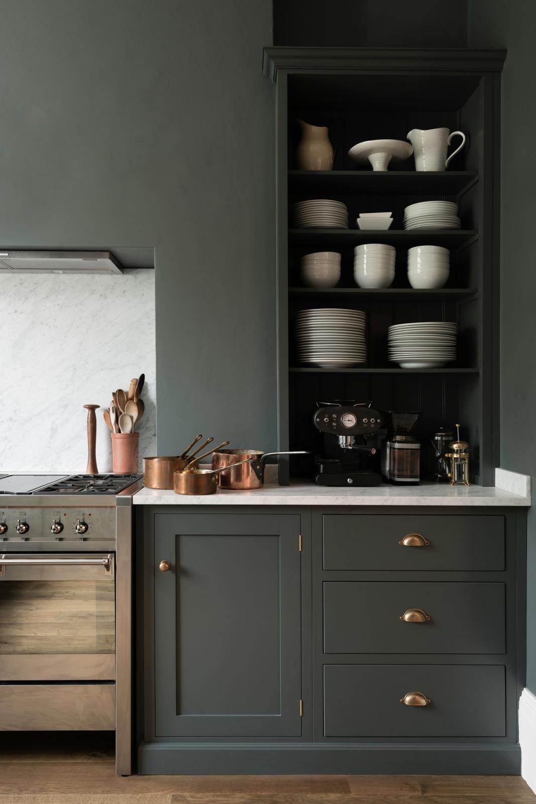 A quiet corner of a charcoal-black classic Shaker kitchen features a U-shaped sink and cooker run with a Carrara marble worktop, matching splashback, and open countertop cupboards. Beautifully decorated with copper pans, simple white ceramic plates and bowls, and a black espresso maker, the family kitchen strikes a wonderful balance between modern style and timeless character.