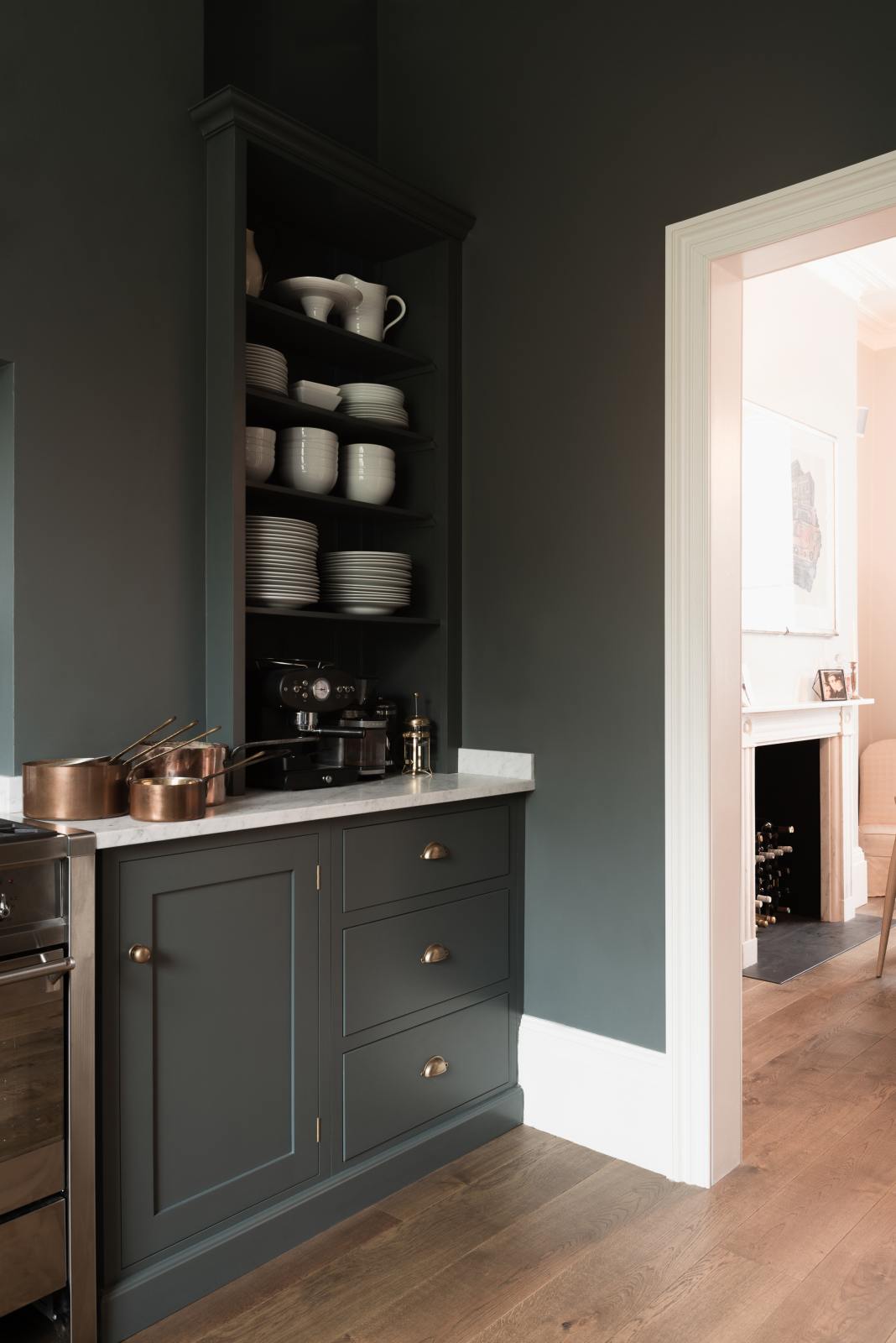 A quiet corner of a charcoal-black classic Shaker kitchen features a U-shaped sink and cooker run with a Carrara marble worktop, matching splashback, and open countertop cupboards. Beautifully decorated with copper pans, simple white ceramic plates and bowls, and a black espresso maker, the family kitchen strikes a wonderful balance between modern style and timeless character.