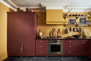 A busy and vibrant Refectory Red Shaker kitchen features an L-shaped sink and cooker run with a Belgian blue limestone worktop, a freestanding upright cupboard, and a classic Shaker canopy. Adorned with vintage items, beloved ceramics, still-life paintings, wooden chopping boards, red lettuce and deep red pomegranates, it creates a bright and wonderfully social kitchen space.
