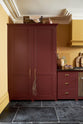 A busy and vibrant Refectory Red Shaker kitchen features an L-shaped sink and cooker run with a Belgian blue limestone worktop, a freestanding upright cupboard, and a classic Shaker canopy. Adorned with vintage items, beloved ceramics, a still-life painting, wooden chopping boards, and red lettuce, creates a bright and wonderfully social kitchen space.