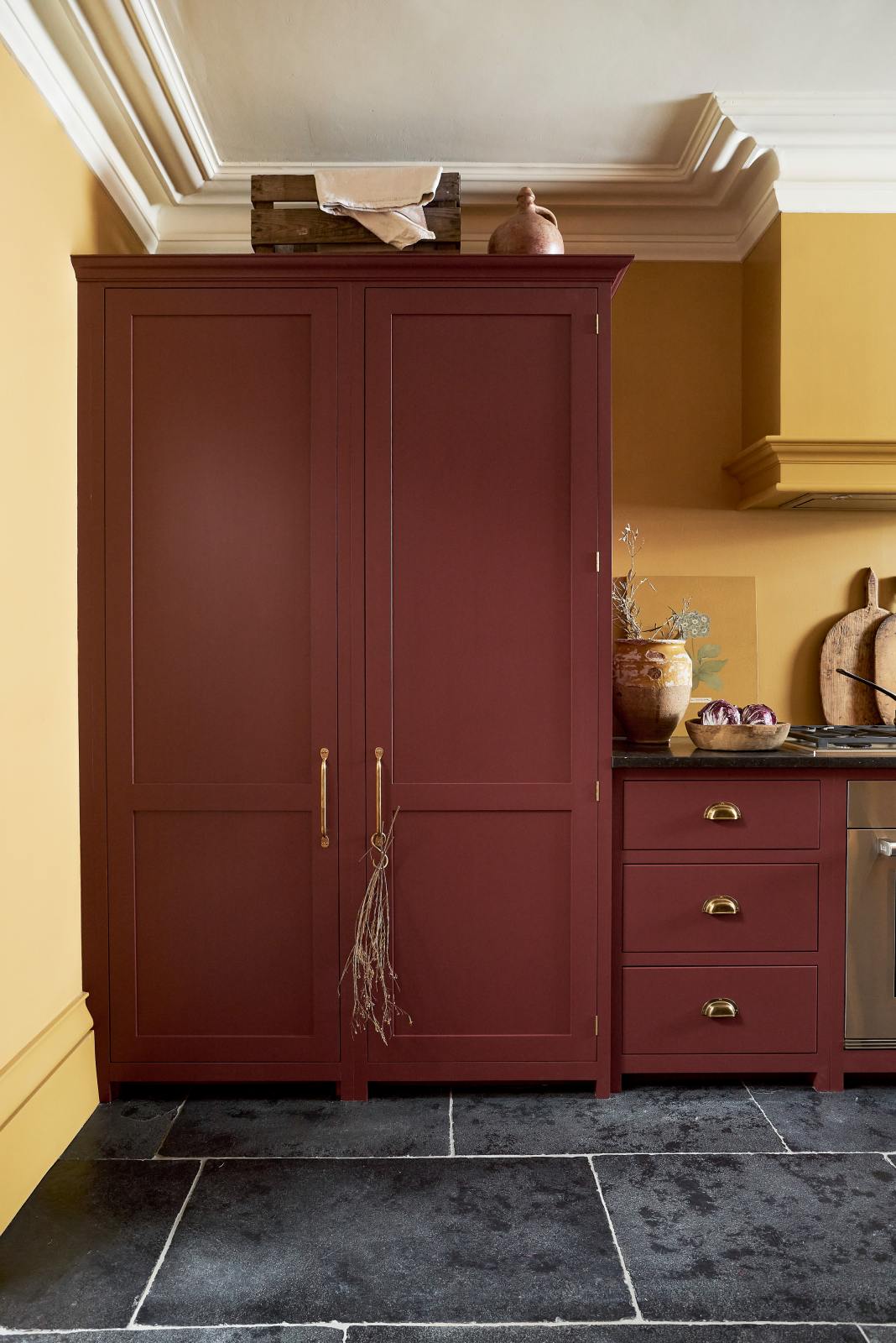 A busy and vibrant Refectory Red Shaker kitchen features an L-shaped sink and cooker run with a Belgian blue limestone worktop, a freestanding upright cupboard, and a classic Shaker canopy. Adorned with vintage items, beloved ceramics, a still-life painting, wooden chopping boards, and red lettuce, creates a bright and wonderfully social kitchen space.