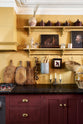 A busy and vibrant Refectory Red Shaker kitchen features an L-shaped sink and cooker run with a Belgian blue limestone worktop, shelves with brass rails, and a classic Shaker canopy. Adorned with vintage items, beloved ceramics, a still-life painting, wooden chopping boards, and dried chillies, creates a bright and wonderfully social kitchen space.