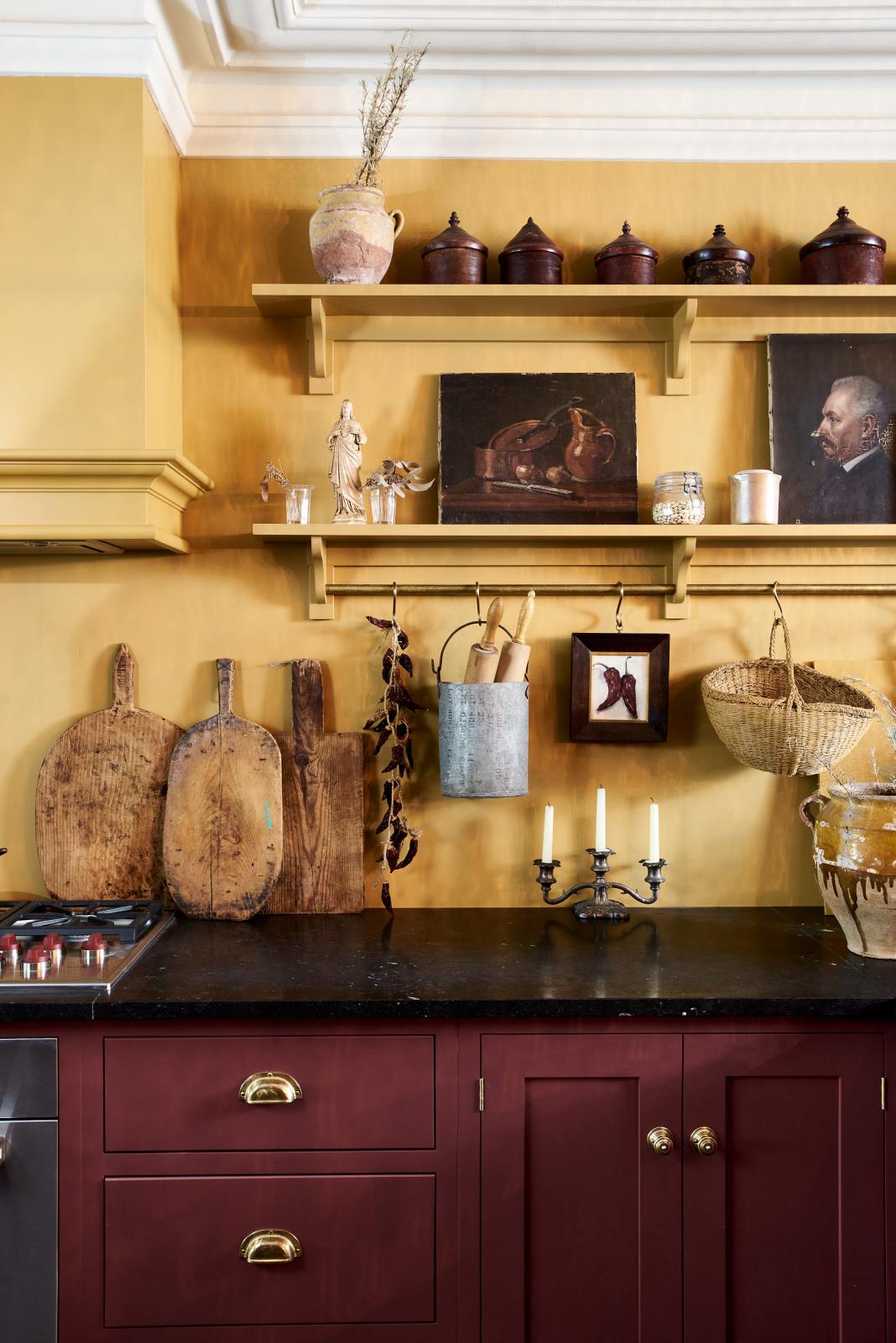 A busy and vibrant Refectory Red Shaker kitchen features an L-shaped sink and cooker run with a Belgian blue limestone worktop, shelves with brass rails, and a classic Shaker canopy. Adorned with vintage items, beloved ceramics, a still-life painting, wooden chopping boards, and dried chillies, creates a bright and wonderfully social kitchen space.