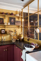 The edge of a busy and vibrant Refectory Red Shaker kitchen features an L-shaped sink and cooker run with a Belgian blue limestone worktop, shelves with brass rails, and a single fluted Belfast sink. Adorned with vintage items, beloved ceramics, a still-life painting, and red lettuce, creates a bright and wonderfully social kitchen space.