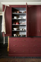 A busy and vibrant Refectory Red Shaker kitchen features a glazed larder with drawers and a Belgian blue limestone worktop. Adorned with vintage items, beloved ceramics, tea towels and bulbs of garlic, creates a bright and wonderfully social kitchen space.