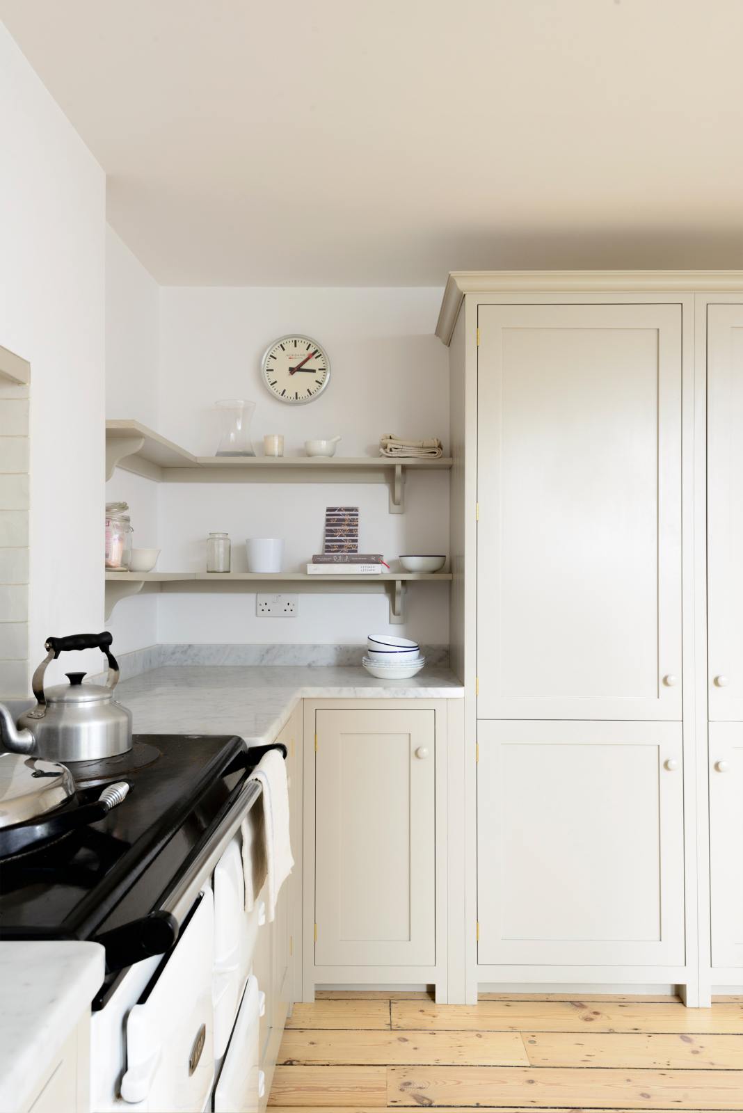 A minimalist, mushroom-coloured shaker kitchen includes a cooker around a chimney breast, a white gas stove, a porcelain splashback, simple shelves and a large pantry cupboard.