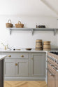 A corner of a spacious and contemporary stone grey Shaker kitchen features an L-shaped sink and cooker run with a Carrara worktop, and a kitchen island. Elegantly decorated with a small bowl of eggs, vintage baskets, artisan ceramics, and books, it creates a cool and inviting space.