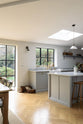An oblique view of a spacious and contemporary stone grey Shaker kitchen features an L-shaped sink and cooker run with a Carrara worktop, and a kitchen island. Elegantly decorated with glass-frilled pendant lights, vintage baskets, artisan ceramics, and deVOL bum stools, it creates a cool and inviting space.
