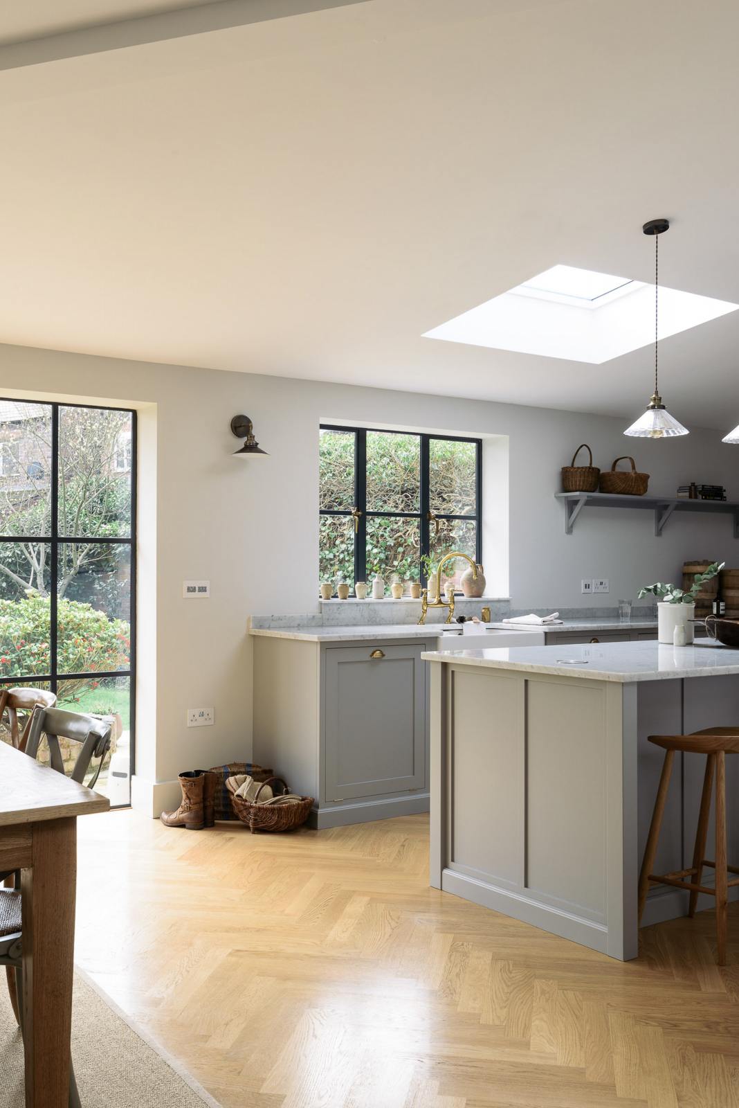 An oblique view of a spacious and contemporary stone grey Shaker kitchen features an L-shaped sink and cooker run with a Carrara worktop, and a kitchen island. Elegantly decorated with glass-frilled pendant lights, vintage baskets, artisan ceramics, and deVOL bum stools, it creates a cool and inviting space.