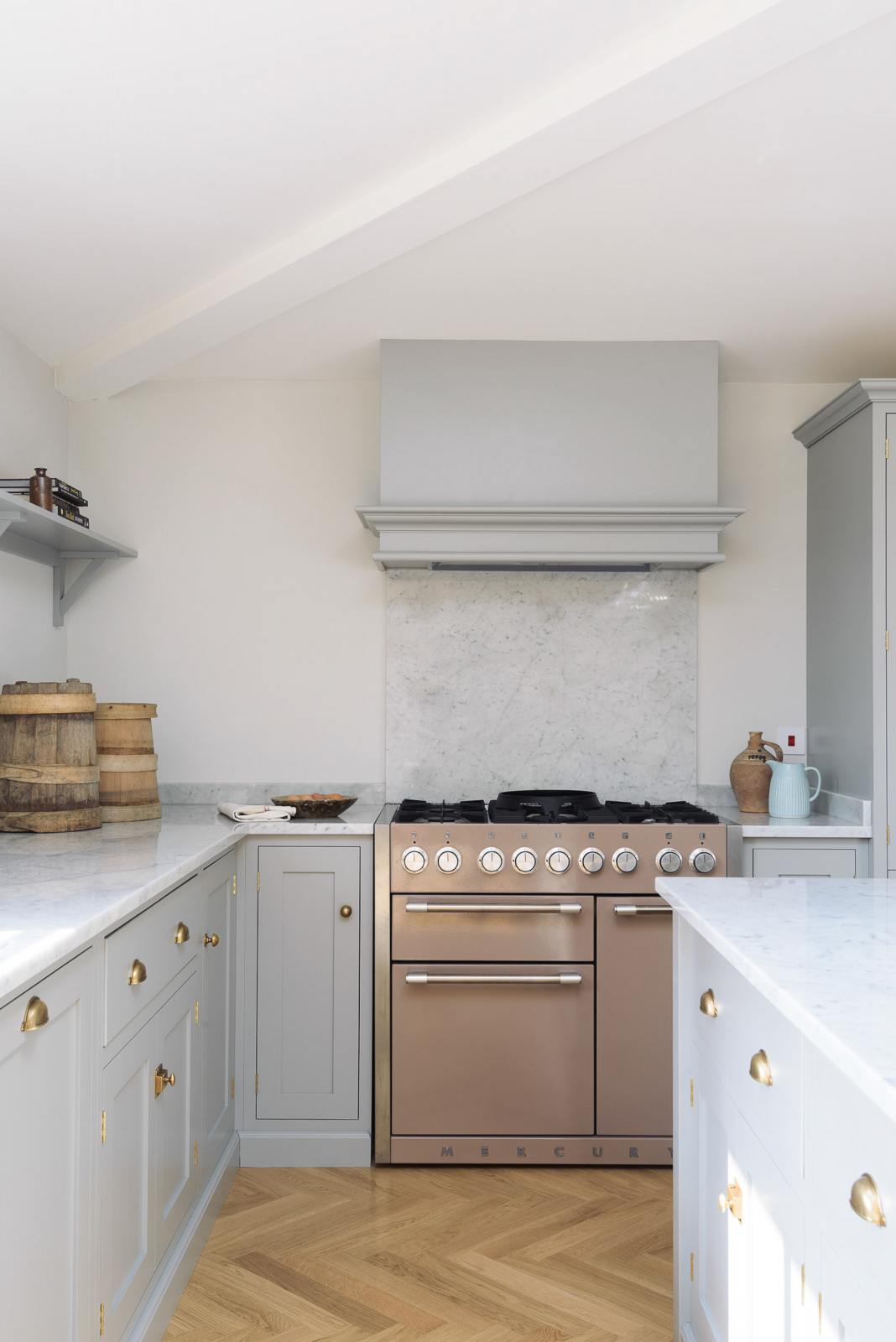 A corner of a spacious and contemporary stone grey Shaker kitchen features an L-shaped sink and cooker run with a Carrara worktop and splashback, a canopy, and a kitchen island. Elegantly decorated with artisan ceramics, a bowl of fresh eggs, and vintage baskets creates a cool and inviting space.
