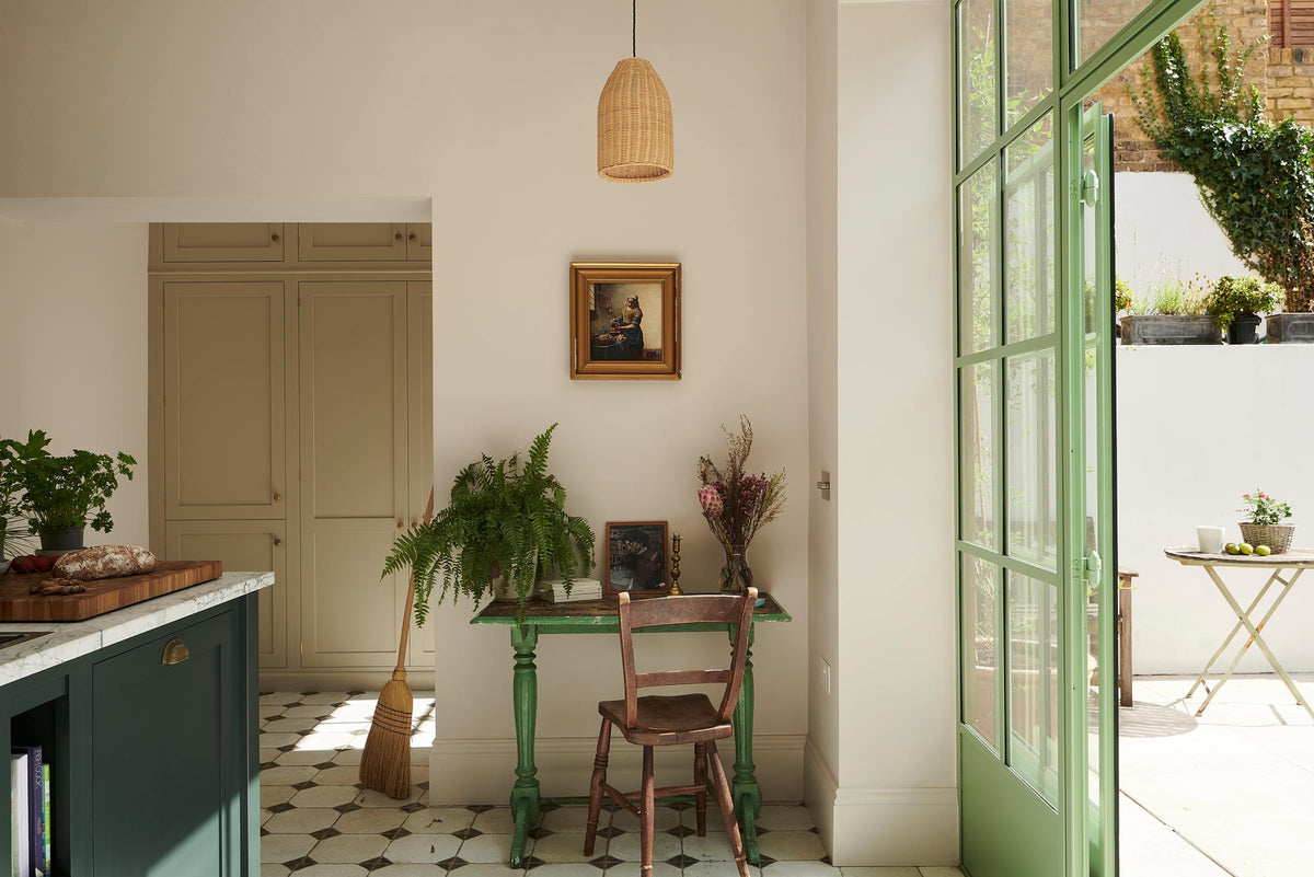 A cosy corner of a Bakehouse Green shaker kitchen features a vintage green wooden desk, a brown chair, and a small painting in a golden frame. This nook also showcases a section of the kitchen island with a Carrara marble worktop, overlooking the pathway to the spacious Mushroom shaker storage and pantry.