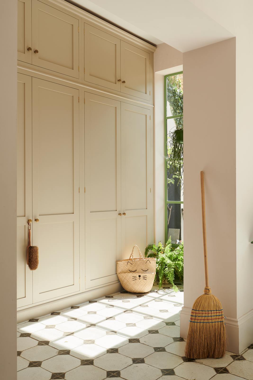 An angled view of the pathway to the spacious, floor-to-ceiling Mushroom shaker storage and pantry. The space features a reclaimed octagonal marble-tiled floor adorned with handmade accessories like a woven basket, broom, and wooden scrubbing brush.