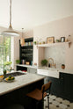 An angled view of the Bakehouse Green shaker kitchen’s cooker kitchen island and sink run features a marble worktop, open cabinets and shelves, along with a double Belfast sink, an aged brass Ionian tap, and a Carrara marble splashback.