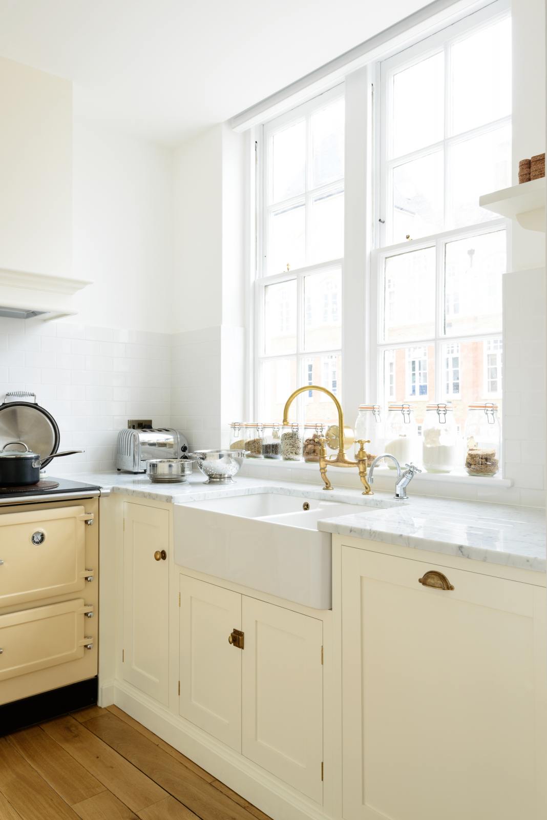 A close-up and angled view of a white linen shaker kitchen sink run featuring a double Belfast sink, an aged brass Ionian tap, and a Carrara marble worktop overlooking a glass window with a cityscape view.