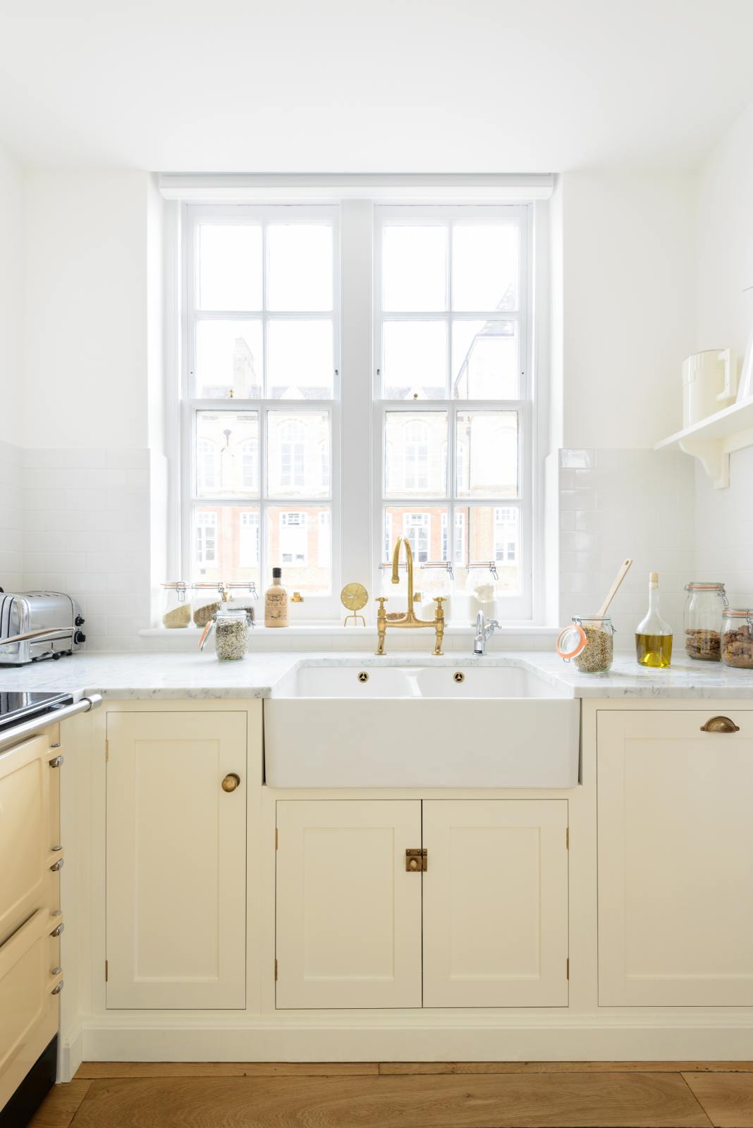A close-up view of a small, white linen shaker kitchen sink run featuring a double Belfast sink, an aged brass Ionian tap, and a Carrara marble worktop, overlooking a glass window with a cityscape view.