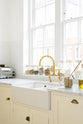 A close-up and angled view of a white linen shaker kitchen sink run featuring a double Belfast sink, an aged brass Ionian tap, and a Carrara marble worktop, overlooking a glass window with a cityscape view.