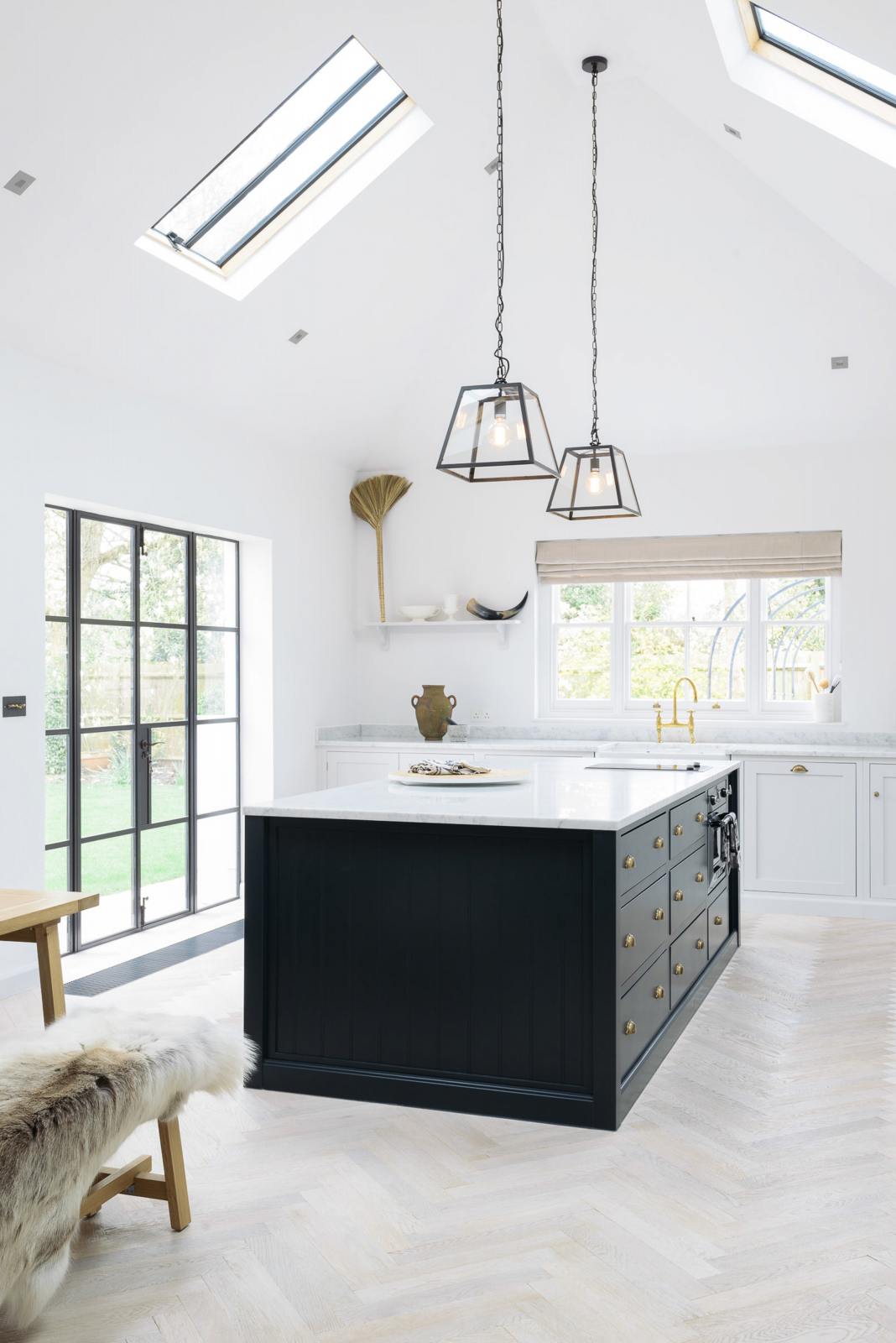 An oblique view of a spacious and artisan Shaker kitchen features an off-white L-shaped sink run with a double Belfast sink and an aged brass Ionian tap, a large pantry-blue kitchen island with a Carrara marble worktop, and a shelf. Cleverly decorated with steel-framed hanging lights, vintage ceramics, and eclectic items serving as artistic displays, it creates a simple yet intriguing kitchen space.