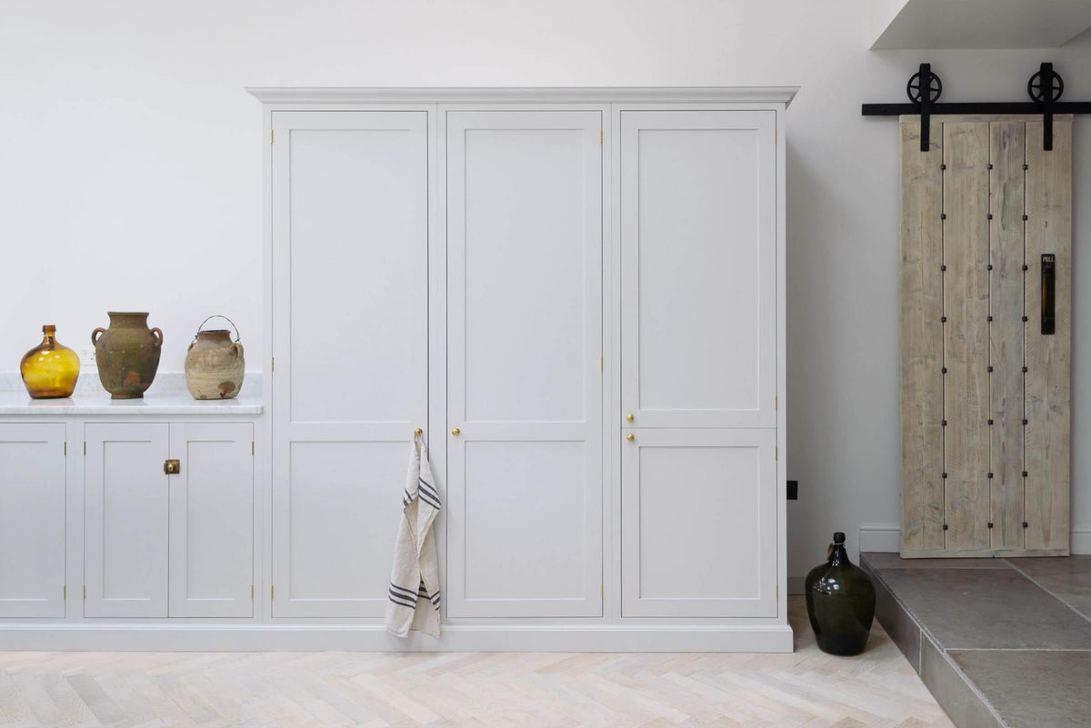 A spacious and artisan Shaker kitchen features an off-white L-shaped sink run with a Carrara marble worktop, a large pantry-blue kitchen island, and a bank of full-height cupboards. Cleverly decorated with vintage ceramics serving as artistic displays, it creates a simple yet intriguing kitchen space.