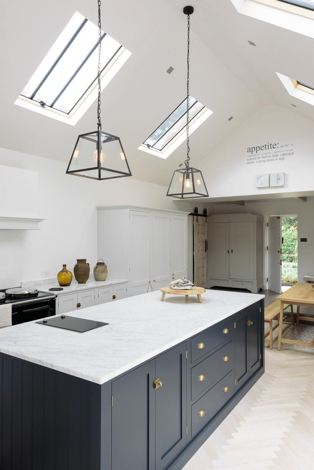 An oblique view of a spacious and artisan Shaker kitchen features an off-white L-shaped sink run with a double Belfast sink and an aged brass Ionian tap, a pantry-blue kitchen island with a Carrara marble worktop, a canopy, and a bank of cupboards. Cleverly decorated with steel-framed hanging lights, vintage ceramics, and the original barn door serving as artistic displays, it creates a simple yet intriguing kitchen space.