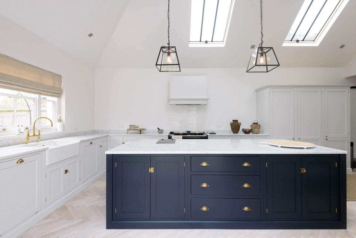 A spacious and artisan Shaker kitchen features an off-white L-shaped sink run with a double Belfast sink and an aged brass Ionian tap, a large pantry-blue kitchen island with a Carrara marble worktop, a canopy, white porcelain splashback tiles, and a bank of cupboards. Cleverly decorated with steel-framed hanging lights, vintage ceramics, and eclectic items serving as artistic displays, it creates a simple yet intriguing kitchen space.
