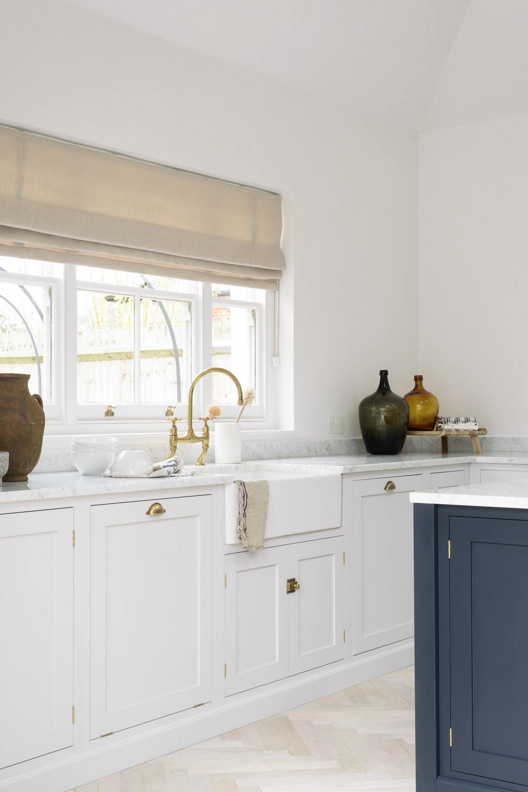 An oblique view of a spacious and artisan Shaker kitchen features an off-white L-shaped sink run with a double Belfast sink, an aged brass Ionian tap, and a pantry-blue kitchen island with a Carrara marble worktop. Cleverly decorated with steel-framed hanging lights, large glasses with narrow necks, and vintage ceramics serving as artistic displays, it creates a simple yet intriguing kitchen space.