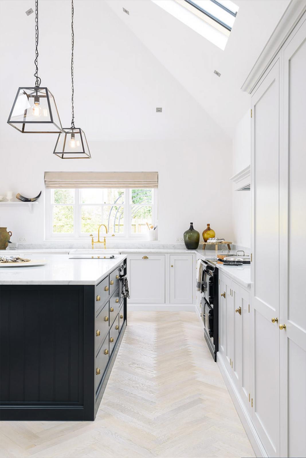 Nestled between an off-white L-shaped sink run with a double Belfast sink and an aged brass Ionian tap, and a pantry-blue kitchen island with a Carrara marble worktop, is a spacious and artisan Shaker kitchen. Cleverly decorated with steel-framed hanging lights, large glasses with narrow necks, and vintage ceramics serving as artistic displays, it creates a simple yet intriguing kitchen space.