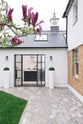 Outside the spacious and artisan Shaker kitchen is the modern and renovated exterior of the Victorian family home, featuring a pristine white wall mixed with the original brick texture, a black tiled roof, a small clock tower, a tiled pathway, freshly cut grass, and a bit of a fuchsia magnolia tree.
