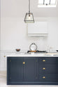 A spacious and artisan Shaker kitchen features an off-white L-shaped sink run, a large pantry-blue kitchen island with a Carrara marble worktop, a canopy, and white porcelain splashback tiles. Cleverly decorated with steel-framed hanging lights, vintage ceramics, and eclectic items serving as artistic displays, it creates a simple yet intriguing kitchen space.