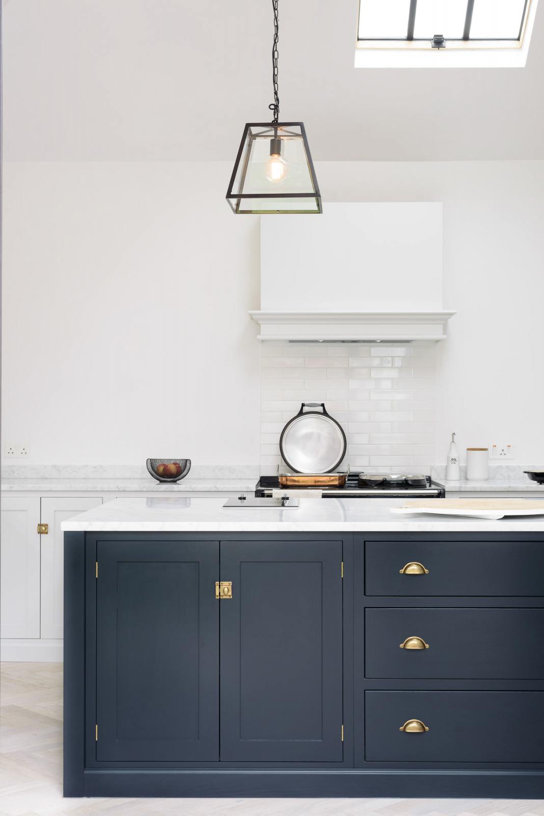 A spacious and artisan Shaker kitchen features an off-white L-shaped sink run, a large pantry-blue kitchen island with a Carrara marble worktop, a canopy, and white porcelain splashback tiles. Cleverly decorated with steel-framed hanging lights, vintage ceramics, and eclectic items serving as artistic displays, it creates a simple yet intriguing kitchen space.
