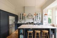 The pantry blue shaker kitchen features creamy Silestone worktops. A stainless steel fridge is built into the cabinetry. Off-white walls frame two pale wall cupboards flanking the chimney breast. The central island features a sink on one side and a breakfast bar with wooden stools on the other, beneath two hanging brass lights.