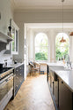The pantry blue shaker kitchen features creamy Silestone worktops. Bright sunshine streams through the tall arched window, illuminating the central island, which houses a sink on one side and a breakfast bar on the other.