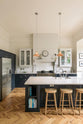 The pantry blue shaker kitchen features creamy Silestone worktops and a built-in stainless steel fridge. Off-white walls frame two pale wall cupboards flanking the chimney breast. The central island holds a sink on one side and a breakfast bar with wooden stools on the other, beneath two hanging brass lights.