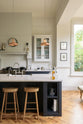 The pantry blue shaker kitchen features creamy Silestone worktops. Off-white walls feature a pale wall cupboard on the chimney breast. The central island holds a sink on one side and a breakfast bar with wooden stools on the other, lit by two brass pendant lights.
