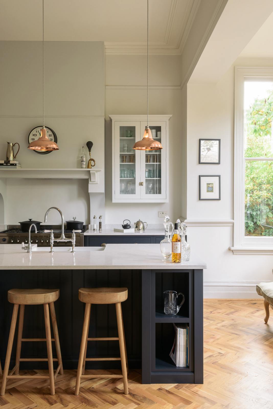 The pantry blue shaker kitchen features creamy Silestone worktops. Off-white walls feature a pale wall cupboard on the chimney breast. The central island holds a sink on one side and a breakfast bar with wooden stools on the other, lit by two brass pendant lights.