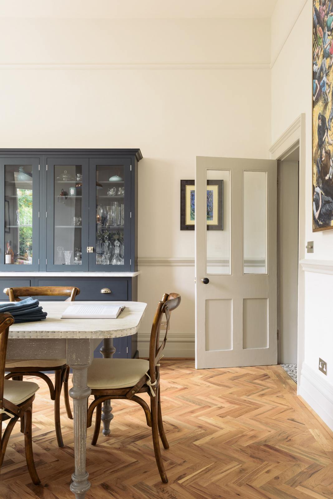 A door is open to welcome people, and a dark navy blue deVOL shaker cupboard stands next to it. In the foreground is a rustic table with vintage wooden chairs pulled up. The room features a herringbone-style wooden flooring and off-white walls. 