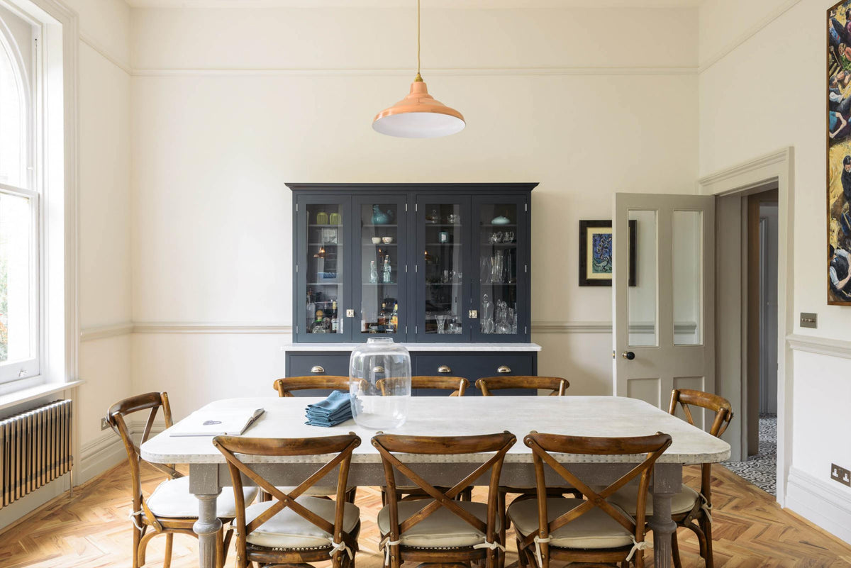 An open door to welcome people, and a dark navy blue deVOL shaker cupboard stands next to it. In the foreground is a huge rustic table with vintage wooden chairs in the middle of the room. A light hangs above the table. The room features a herringbone-style wooden flooring and off-white walls. 