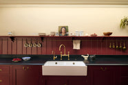 A close-up look at the simple yet elegant Refectory red shaker kitchen features a long L-shaped sink and cooker run with a black granite worktop, a tongue and groove splashback, and a long shelf with brass rails. Beautifully decorated with artisan ceramics, antique champagne glasses, an oil painting, and fresh fruits, creating a bold yet understated kitchen space.