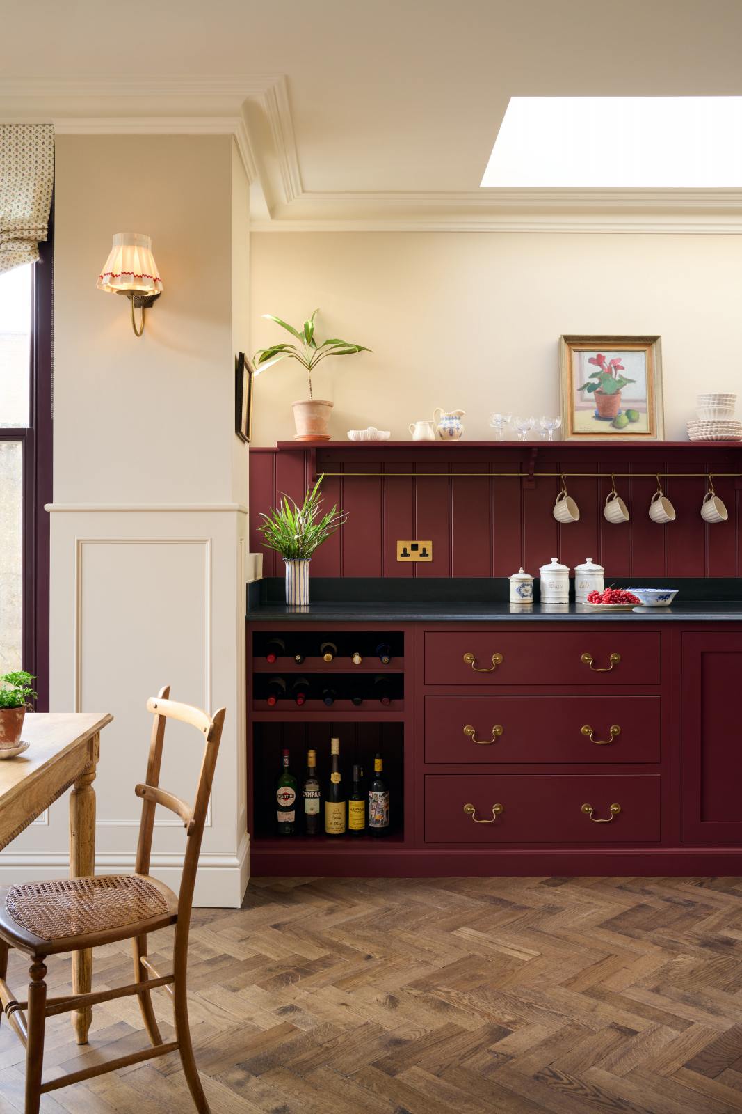 A simple yet elegant Refectory red shaker kitchen features a long L-shaped sink and cooker run with a black granite worktop, a tongue and groove splashback, and a long shelf with brass rails. Beautifully decorated with artisan ceramics, antique champagne glasses, an oil painting, fresh fruits, and wooden parquet flooring, creating a bold yet understated kitchen space.