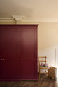 A quiet corner of a simple yet elegant Refectory red shaker kitchen features a bank of pantry cupboards. Beautifully decorated with a wooden chair, red cauliflower, a brown shopping bag, and wooden parquet flooring, creating a bold yet understated kitchen space.