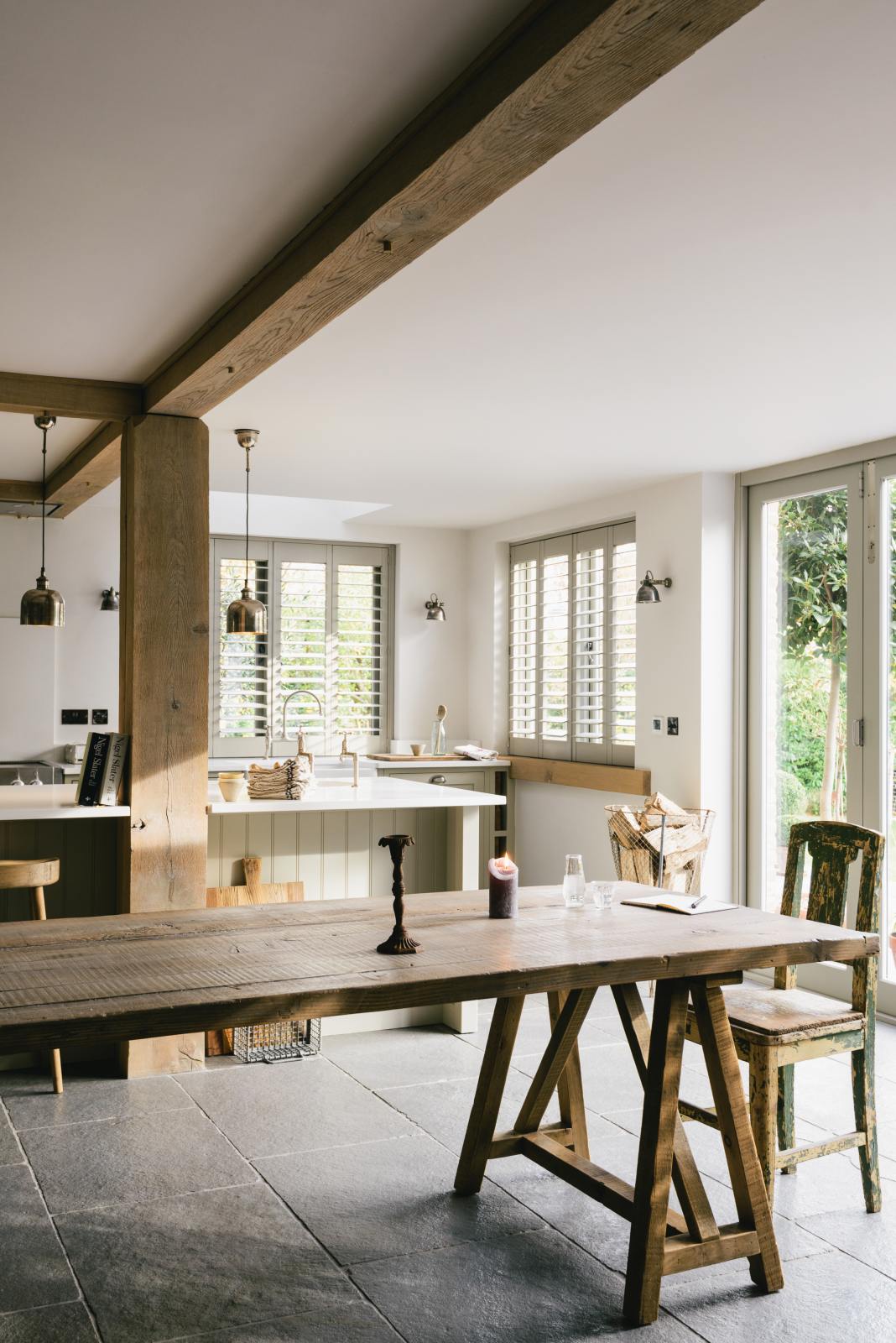 An oblique view of a tranquil and calm Shaker kitchen features a long sink and cooker run with a Silestone worktop and splashback, a single Belfast sink with a nickel Ionian tap, and a long kitchen island with a breakfast nook. Beautifully decorated with a vintage dining table that matches the raw wooden beams, silver pendant lights, and a steel firewood basket, this kitchen creates a lovely, simple space that embraces natural materials.