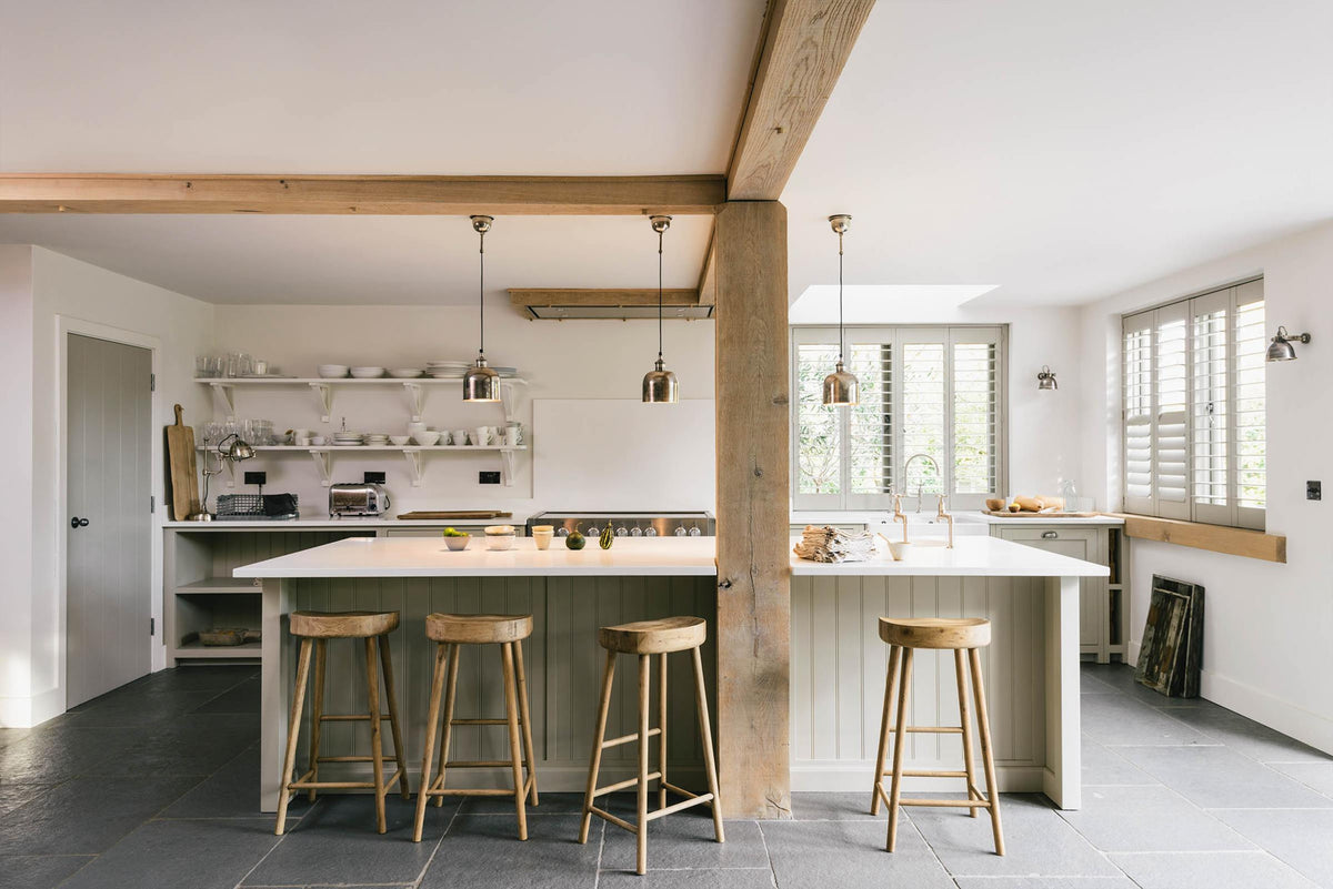 A tranquil and calm Shaker kitchen features a long sink and cooker run with a Silestone worktop and splashback, a single Belfast sink with a nickel Ionian tap, a long kitchen island with a breakfast nook, and shelves. Beautifully decorated with vintage stools that match the raw wooden beams, muted ceramics, silver pendant lights, and a steel firewood basket, this kitchen creates a lovely, simple space that embraces natural materials.